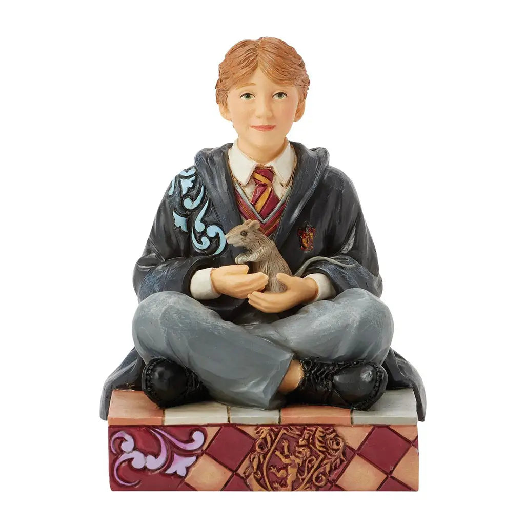 Front view of Jim Shore Ron Weasley figurine, smiling while holding Scabbers the rat, dressed in Gryffindor robes, sitting cross-legged on a decorated base with crest.