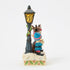 Hand-painted Jim Shore figurine of Rudolph the Red-Nosed Reindeer beside a black lamppost with yellow glow, joined by two elves in blue coats and hats, standing on a holly-trimmed snowy base.