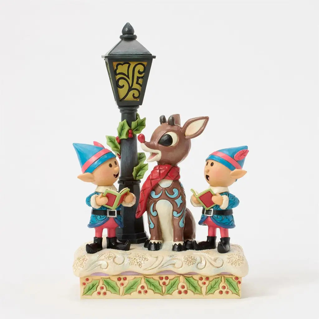 Front view of Jim Shore Rudolph figurine with two elves singing beside a lamppost, detailed with swirling patterns, holly accents, and a snow-covered base with green and red trim.