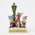 Front view of Jim Shore Rudolph figurine with two elves singing beside a lamppost, detailed with swirling patterns, holly accents, and a snow-covered base with green and red trim.