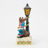 Side angle of Jim Shore Rudolph figurine with lamppost and elves, showing carved details, holly leaves, snowy base, and bright holiday colors in folk art style.