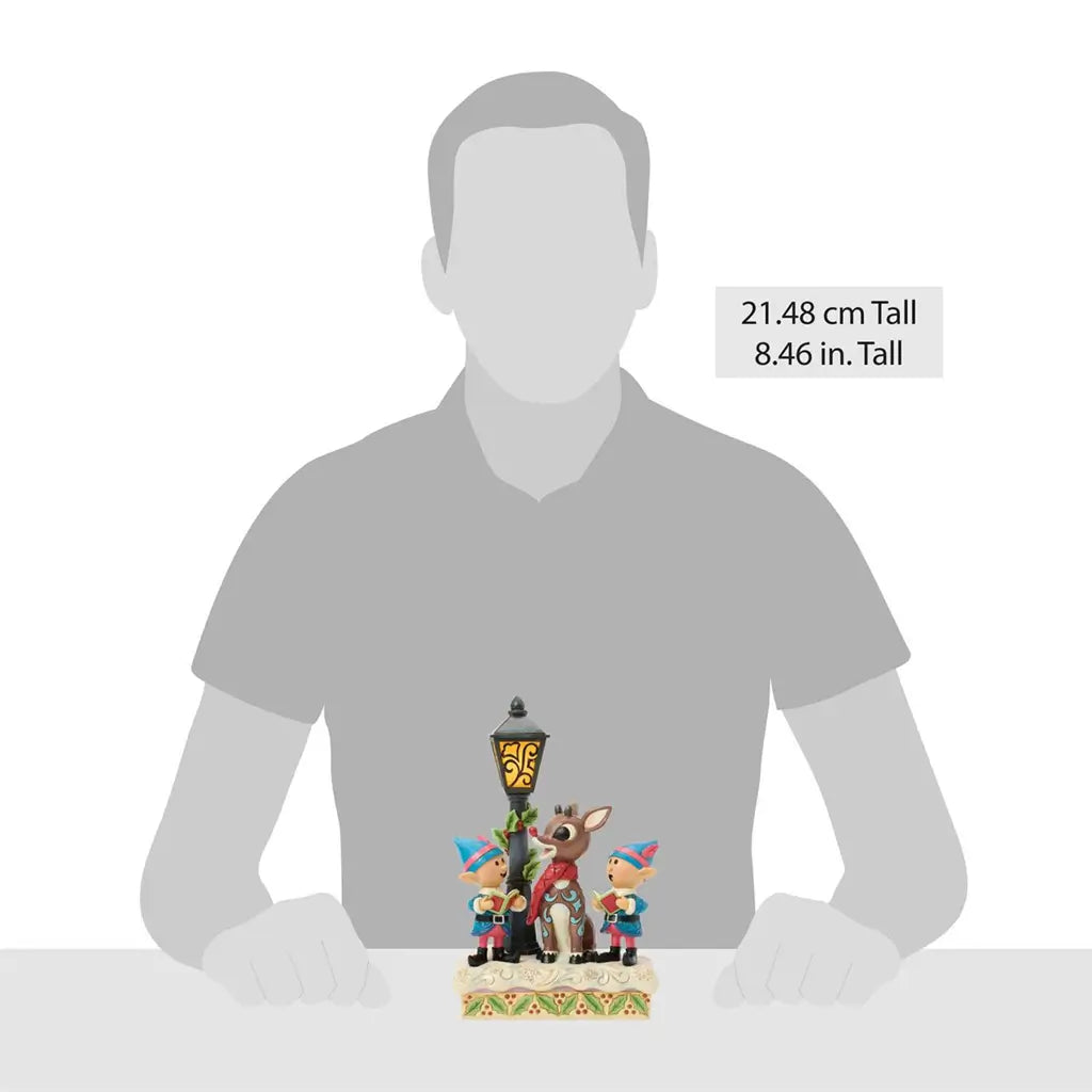 Scale view of Jim Shore Rudolph and elves figurine, showing 8.46-inch tall collectible with lamppost, holly-trimmed snow base, and bright hand-painted holiday colors.
