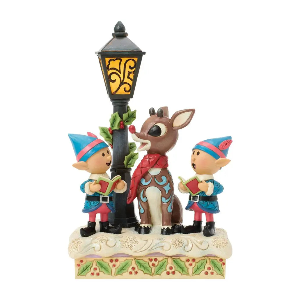 Jim Shore Christmas figurine of Rudolph and elves under a lamppost, hand-carved with folk art patterns, holly garlands, and snowy base, ideal for festive holiday displays.