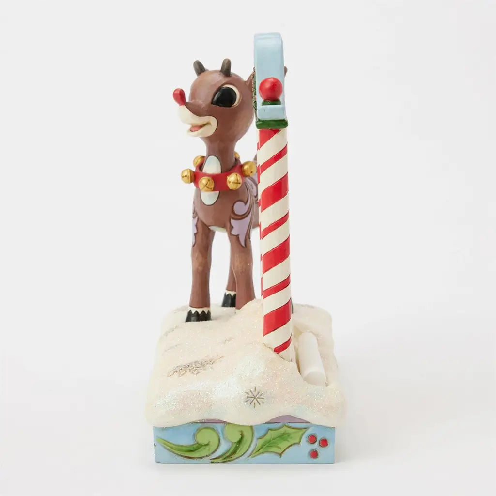Side view of Jim Shore Rudolph countdown figurine, with reindeer facing forward beside candy cane-striped holiday sign on snowy, holly-trimmed base.