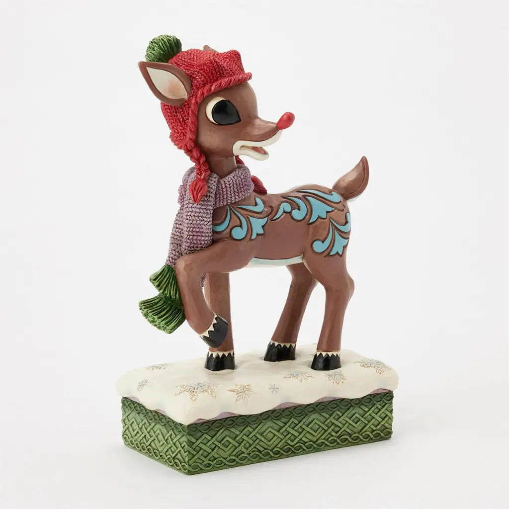 Detailed Rudolph figurine wearing red knit hat, green pom-pom, and purple scarf, standing on snowflake base with textured green edge, angled view from the front side.
