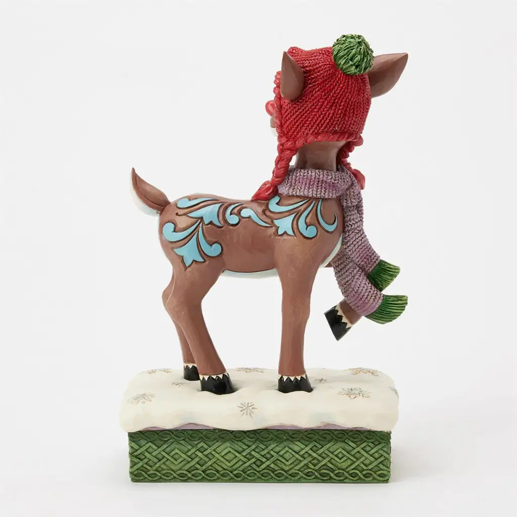 Brown reindeer figurine with ornate blue patterns, red knit hat, and purple scarf, standing on white snowflake base with green woven trim, viewed from the back.