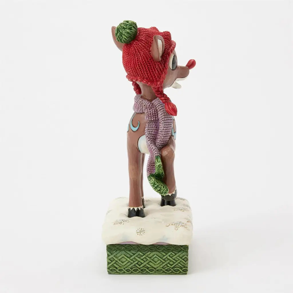 Brown reindeer figurine with red knit hat and green pom-pom, purple scarf, and blue side patterns, standing on snowy base with green trim, viewed from side angle.