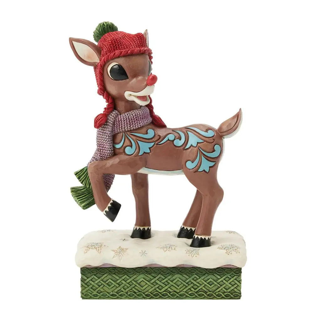 Front view of Rudolph figurine in red knit hat with green pom-pom, purple scarf, and blue side detailing, standing on snowflake-covered base with green textured trim.
