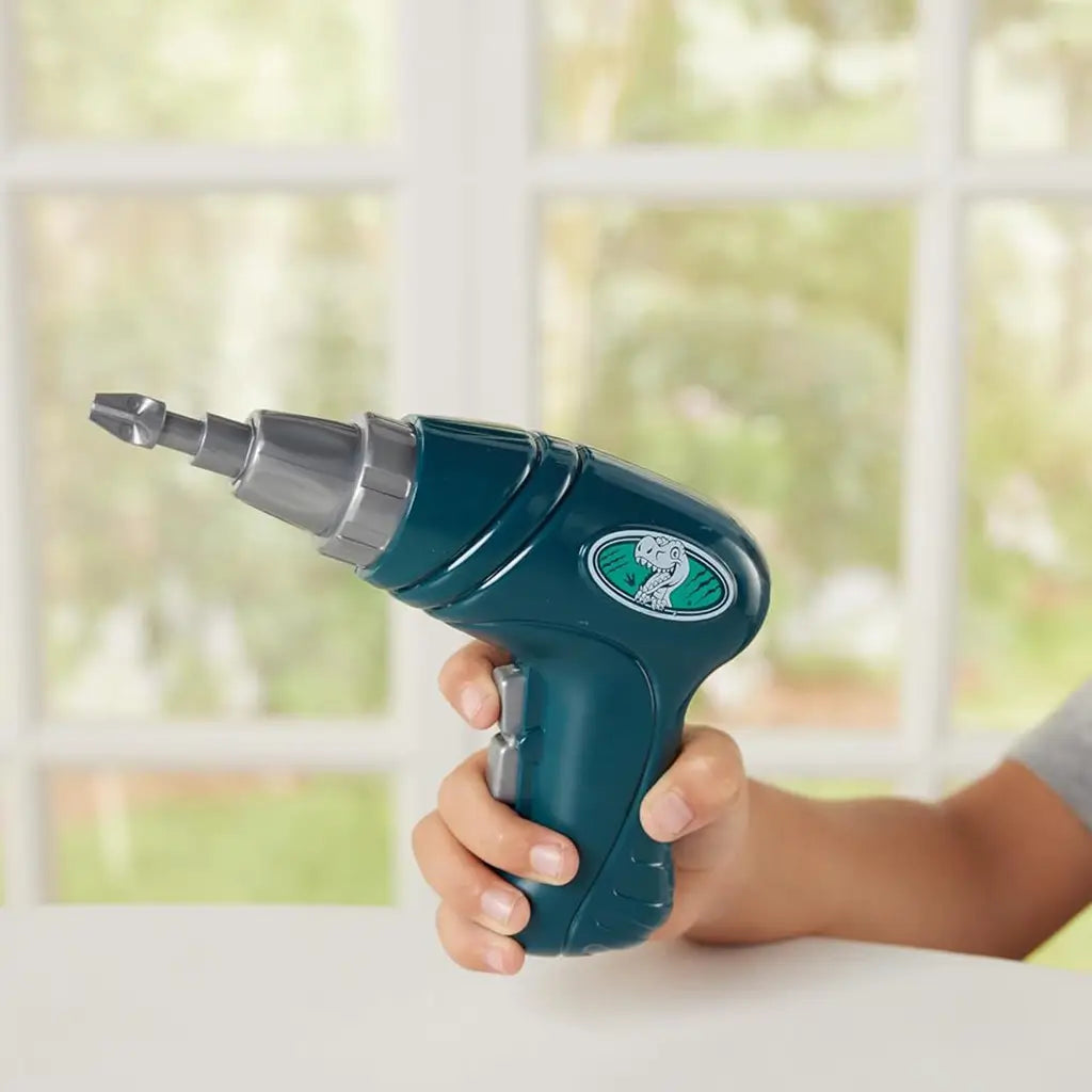Child’s hand holds a teal plastic toy drill with silver interchangeable bit and a dinosaur logo on the handle in front of a softly lit window background.