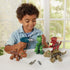 Young boy in a light blue shirt uses a green toy drill to assemble a green plastic dinosaur while three other screw-together dinosaurs sit on a white table in front of a window.