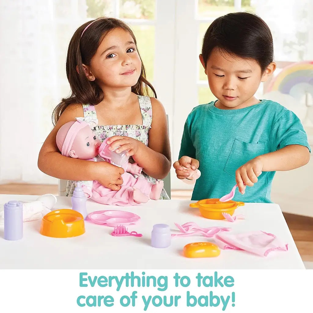 Two children play at a white table with Kidoozie Doll Care Playset, one feeding a baby doll with a bottle while the other uses a spoon and bowl from the included accessories.
