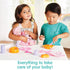 Two children play at a white table with Kidoozie Doll Care Playset, one feeding a baby doll with a bottle while the other uses a spoon and bowl from the included accessories.
