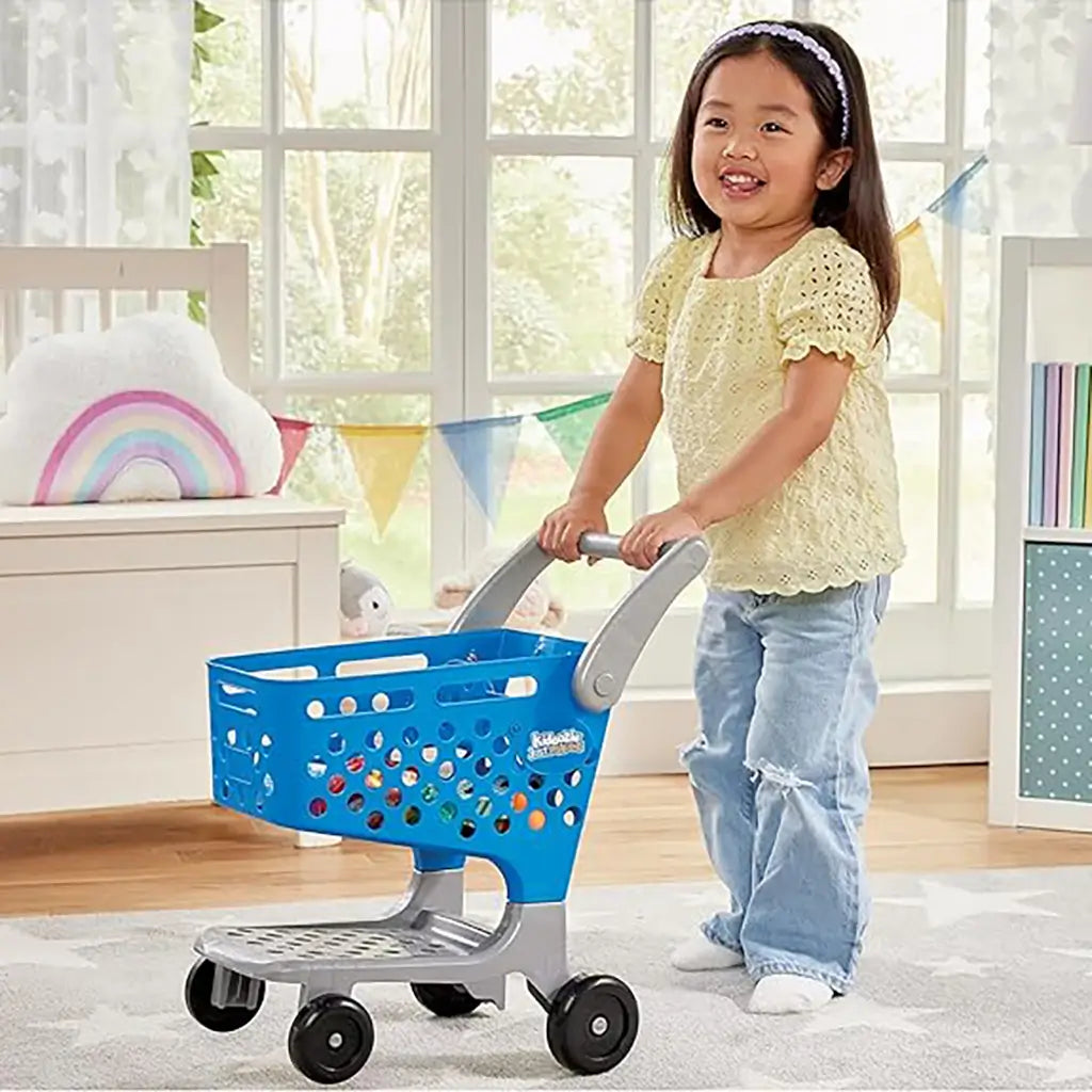 Smiling young girl in a yellow top and jeans pushes a blue Kidoozie toy shopping cart with gray handle inside a playroom decorated with rainbow pillow and festive bunting.