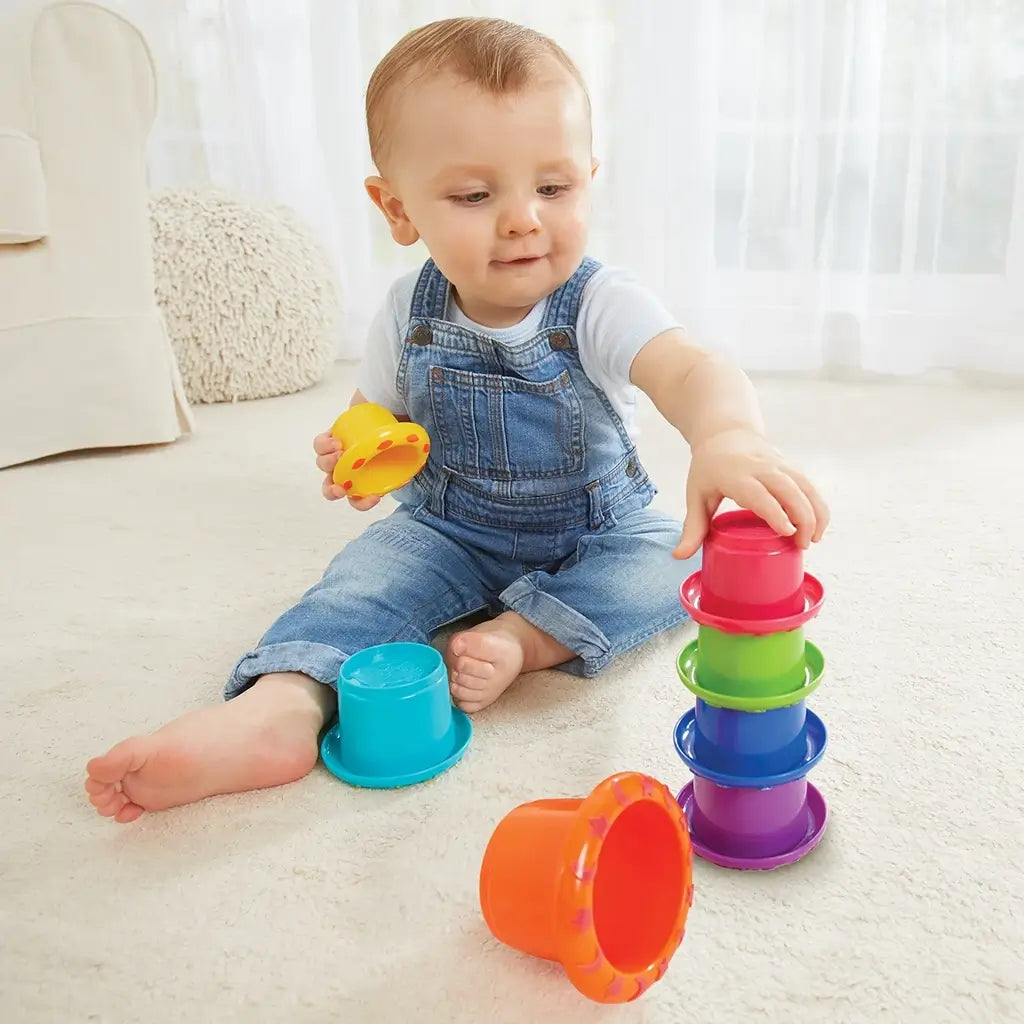 Smiling baby boy in denim overalls sits on beige carpet, stacking colorful Kidoozie cups into a tower while holding a yellow piece, engaging in motor skill and coordination play.