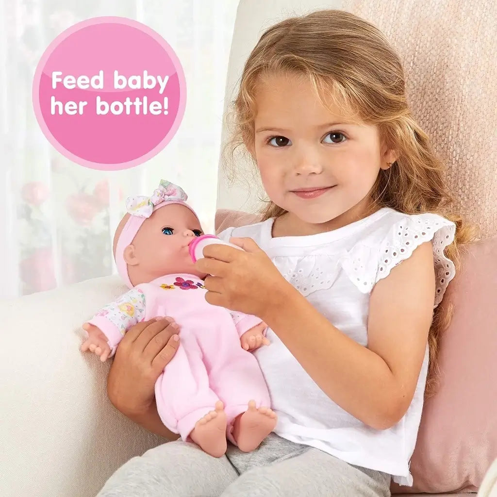 Young girl in a white shirt sits on a chair feeding her Kidoozie Sweetie Doll with a pink baby bottle, demonstrating nurturing playtime.