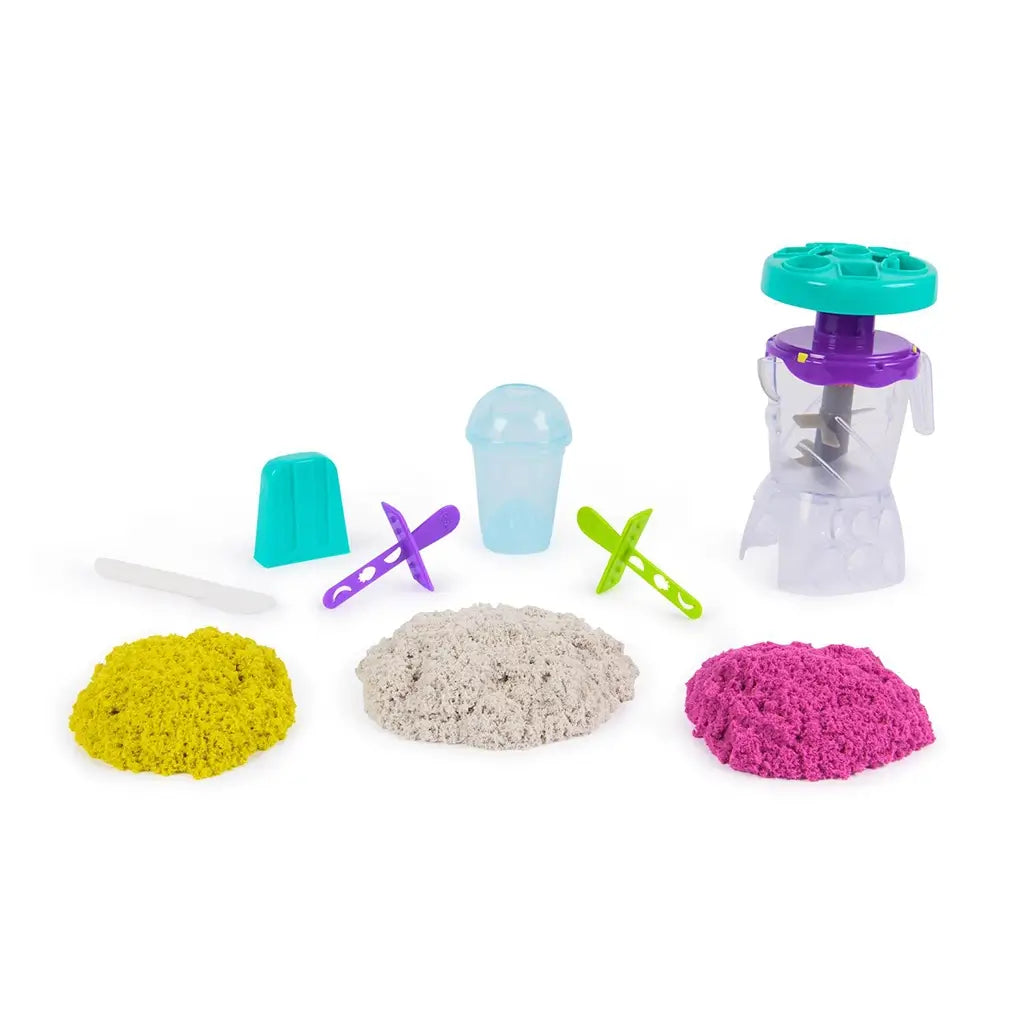 Kinetic Sand Smoothie Mixer playset contents arranged with yellow, white, and pink sand piles, clear cups, popsicle molds, utensils, and blender toy for mix, blend, and mold play on white.