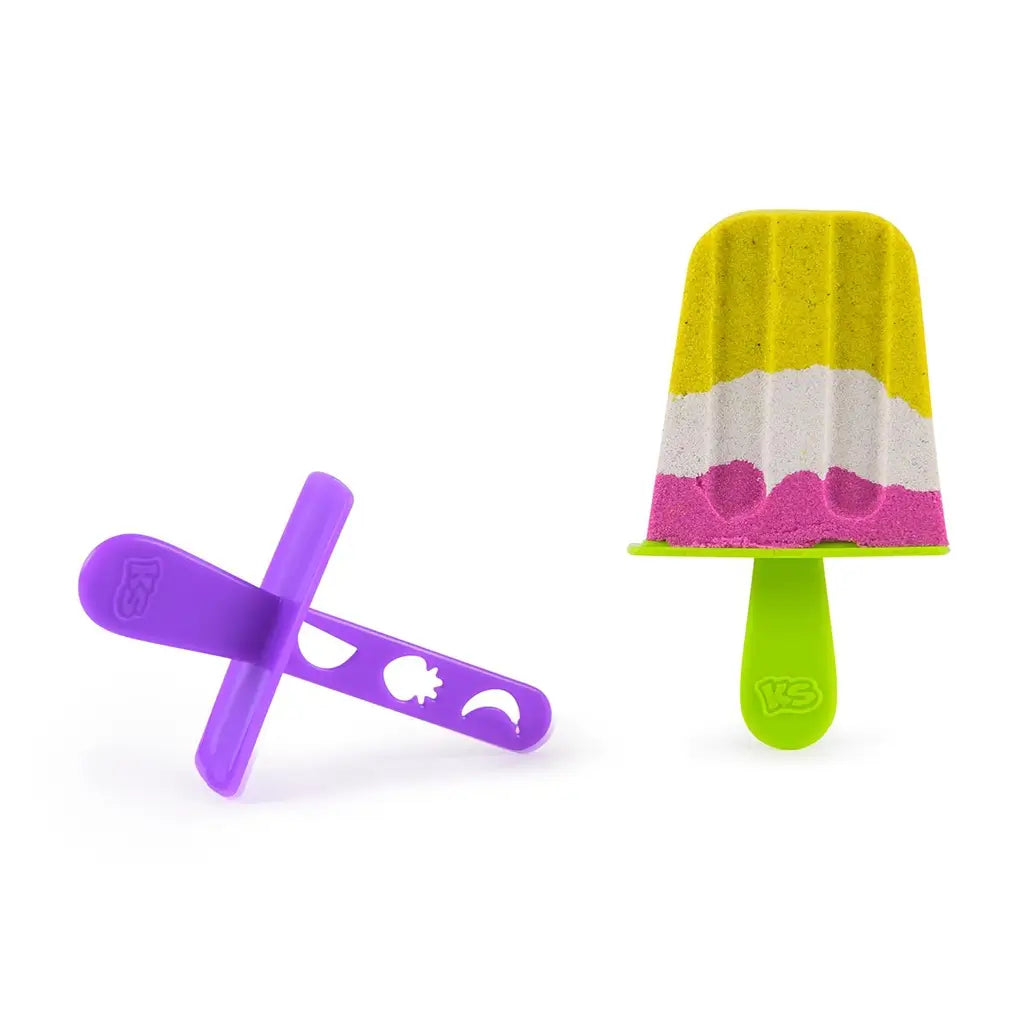 Yellow, white, and pink layered Kinetic Sand popsicle molded with smooth ridges, paired with purple and green plastic stick tools, designed for pretend frozen treat play on white background.
