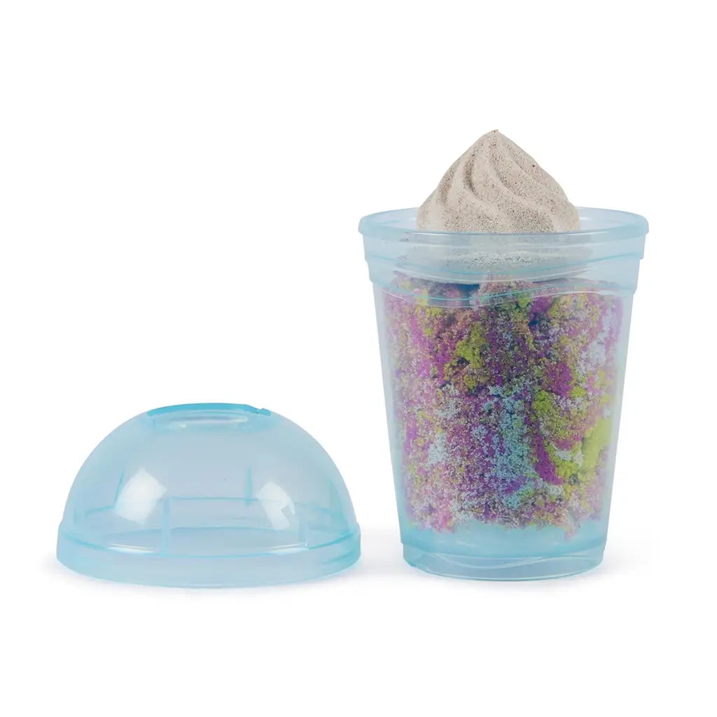 Clear plastic smoothie cup filled with layered yellow, white, pink, and mixed Kinetic Sand topped with molded swirl, showing smooth texture and colorful blending effect on a white background.