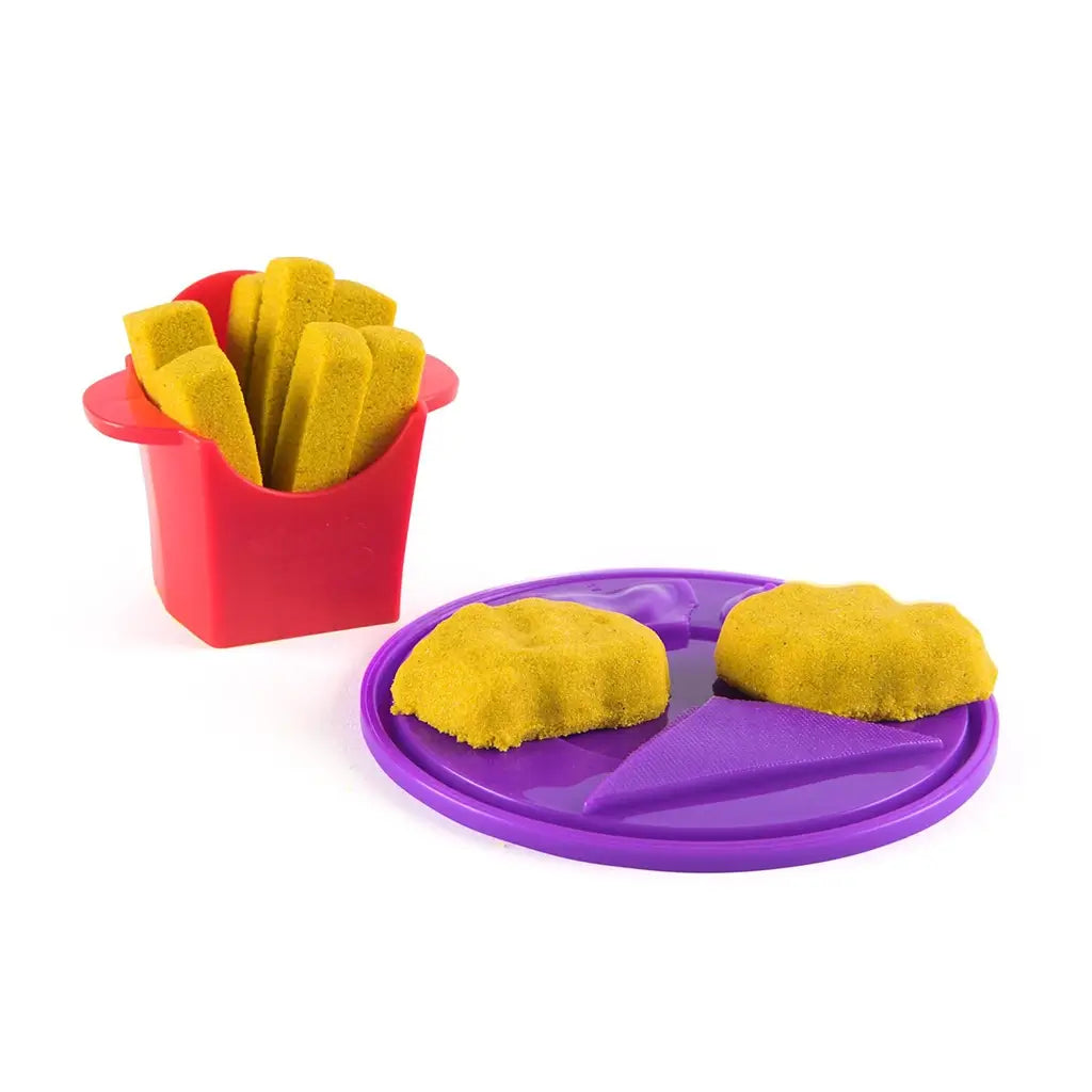 Kinetic sand pretend food set in yellow molded fries and nuggets with soft textured pieces, arranged on purple tray with red container, designed for imaginative play on a white background.