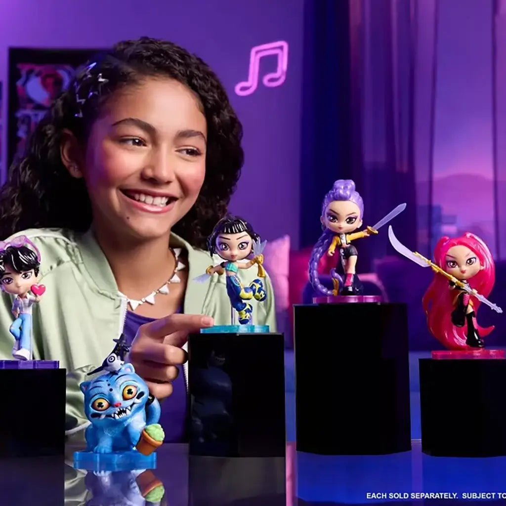 KPop Demon Hunters collectible figures in vivid purple, pink, blue and teal plastic stand on black display blocks as a smiling girl points to the collection in a purple-lit room.