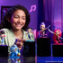 KPop Demon Hunters collectible figures in vivid purple, pink, blue and teal plastic stand on black display blocks as a smiling girl points to the collection in a purple-lit room.