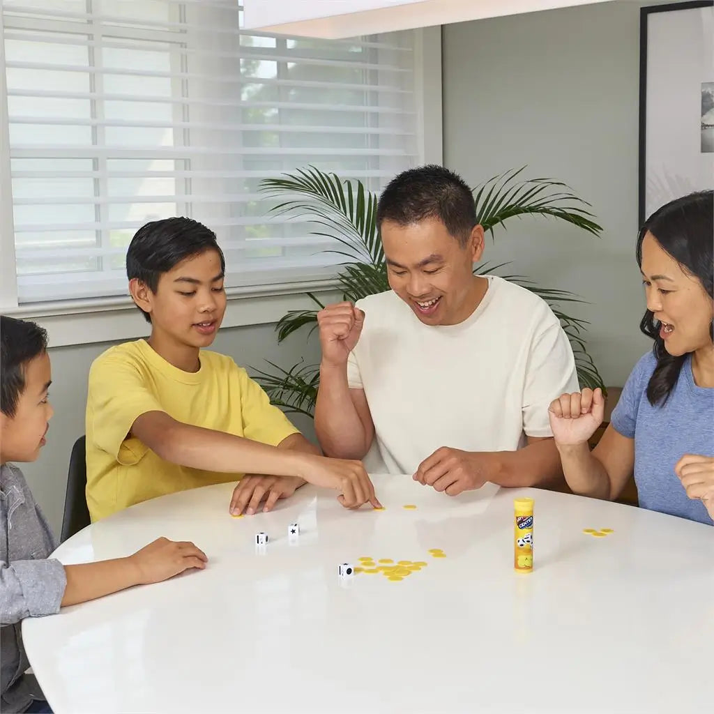 Left Center Right dice game in yellow plastic with round chips and white symbol dice, played by a smiling family seated around a white table in a bright home setting.