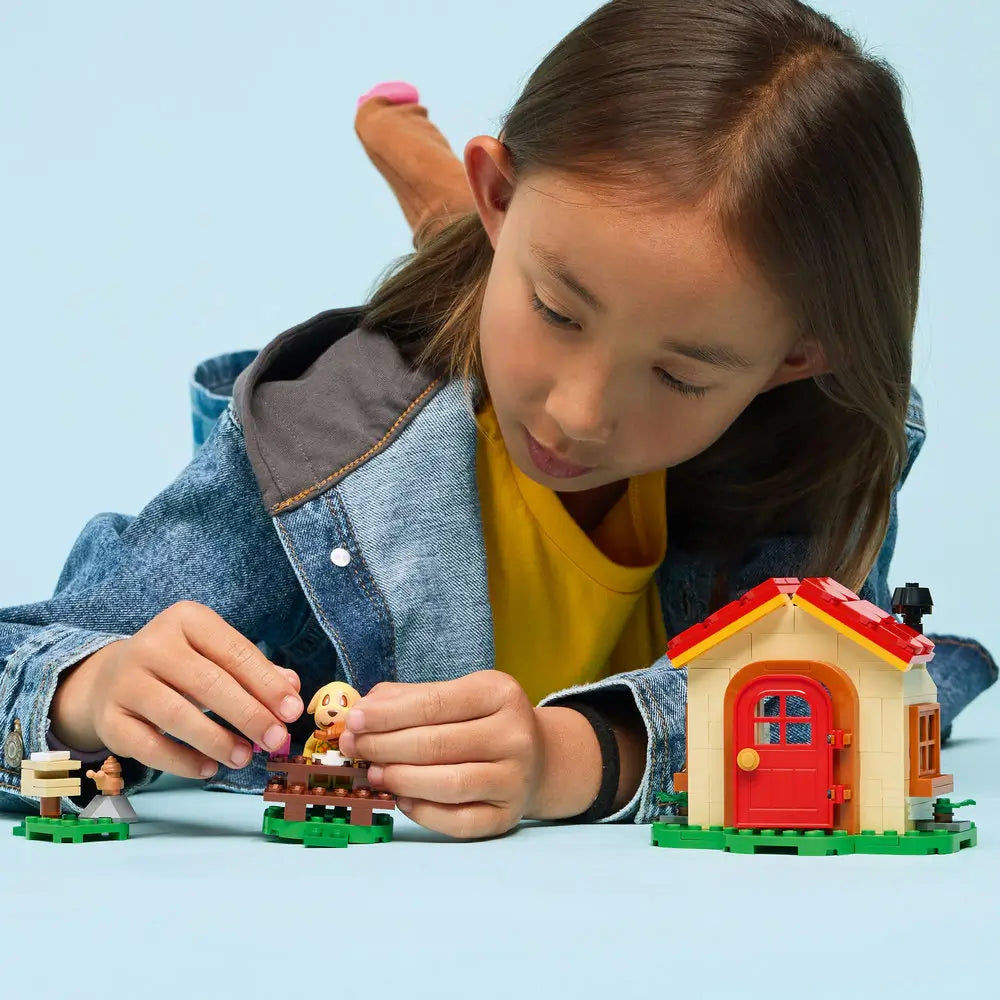 LEGO Animal Crossing Goldie's Cozy House playset features a child playing with the tan cottage, Goldie character and outdoor accessories on a light blue background with detailed LEGO elements and realistic display presentation.
