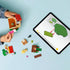 LEGO Animal Crossing Goldie's Cozy House playset shows hands building the cottage with Goldie, accessories and a tablet displaying interactive instructions on a light blue table with detailed LEGO elements and realistic display presentation.