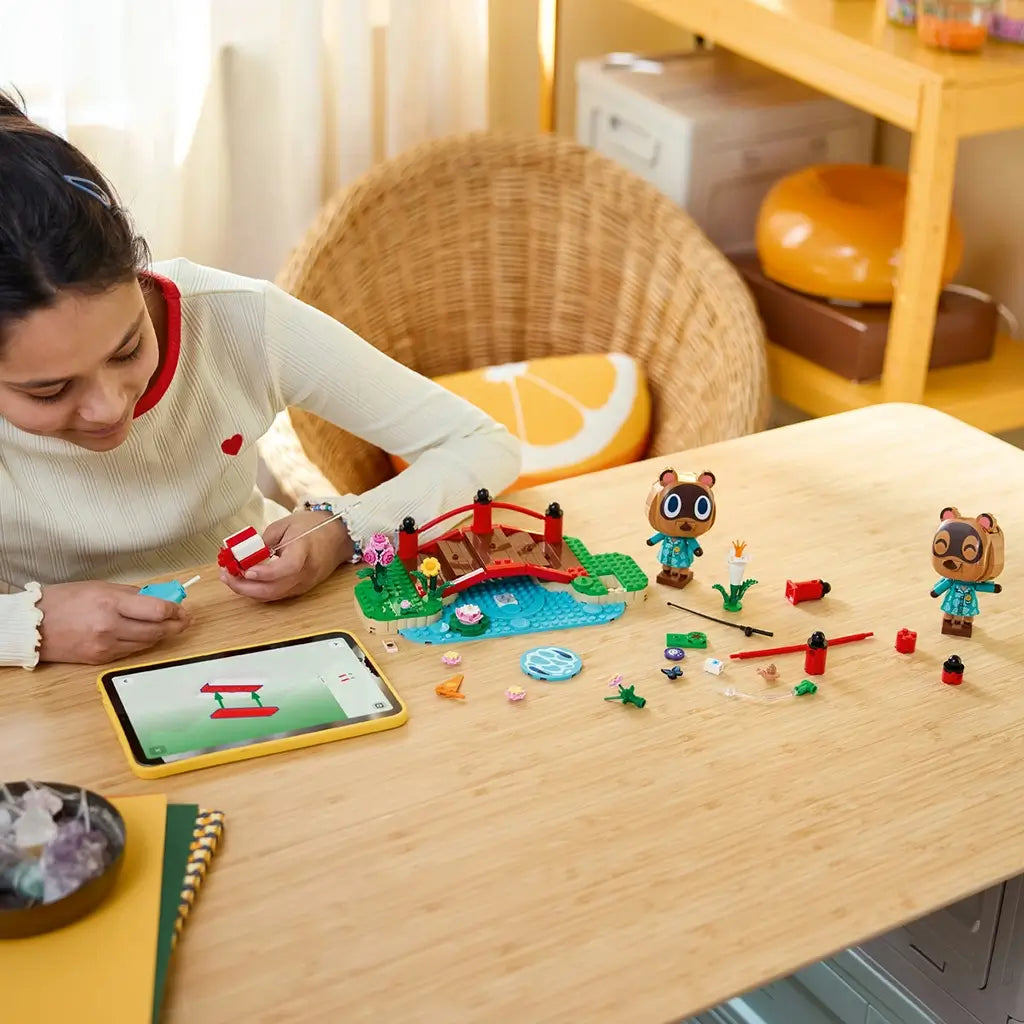 LEGO Animal Crossing Timmy & Tommy playset in red, green, blue, and brown plastic is assembled by a child at a wooden table with loose pieces, two figures, and tablet instructions.