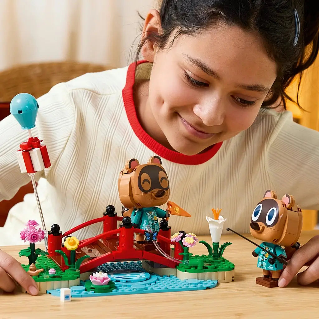 LEGO Animal Crossing Timmy & Tommy playset in red, green, blue, brown, and pink plastic is explored by a child arranging two figures around a flower-lined bridge and stream.