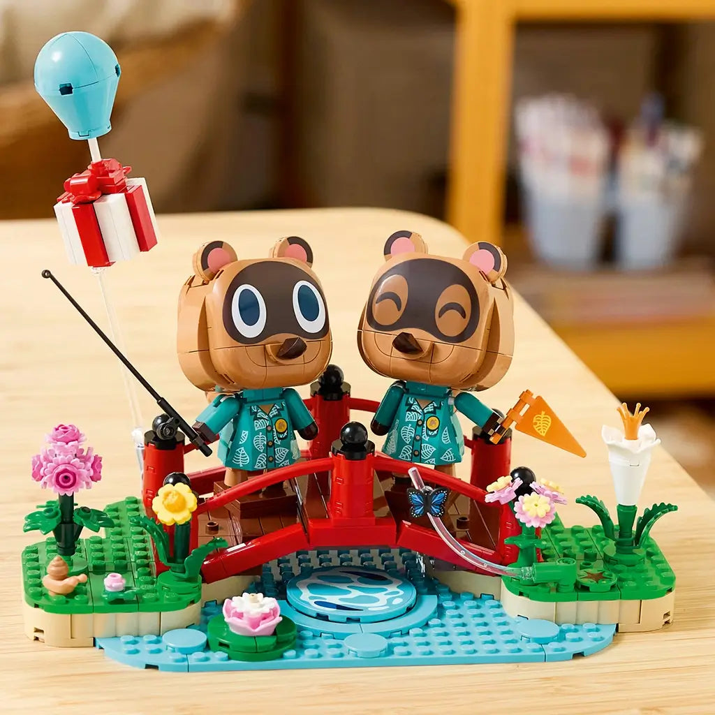 LEGO Animal Crossing Timmy & Tommy playset in red, green, blue, and brown plastic displays two raccoon figures on a curved bridge with flowers, stream, fishing rod, and balloon gift.