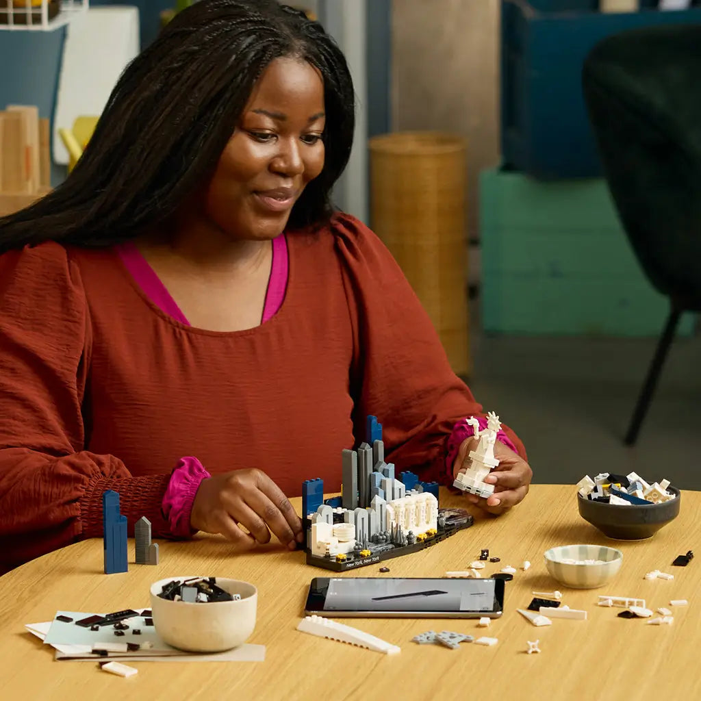 LEGO Architecture New York City building set uses black, white, gray and blue plastic bricks as an adult assembles the skyline model at a table with loose pieces.