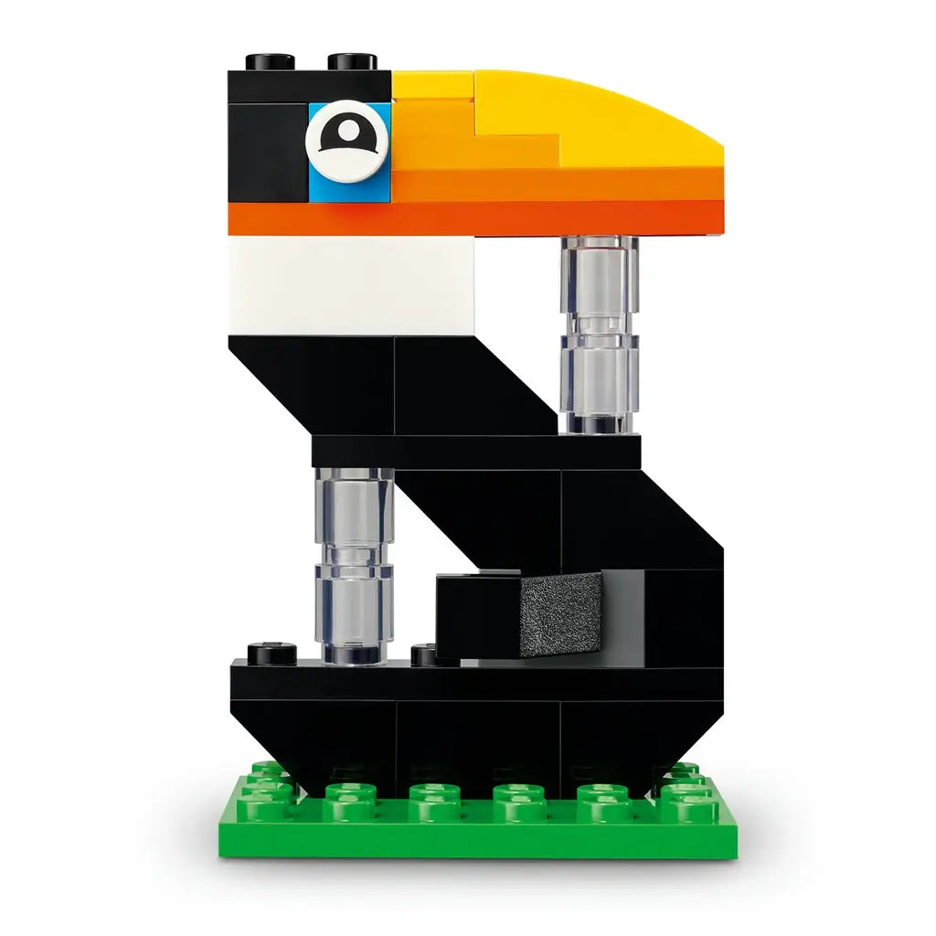 LEGO toucan number display in black, white, yellow and orange plastic bricks, shaped as a stylized bird-themed numeral for birthday decor on a green base against a plain backdrop.