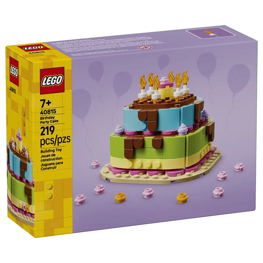 LEGO birthday cake building set featuring yellow glossy cardboard packaging and a colorful plastic brick cake, layered with blue, green and pink frosting details, candles and party accents on a purple studio background.