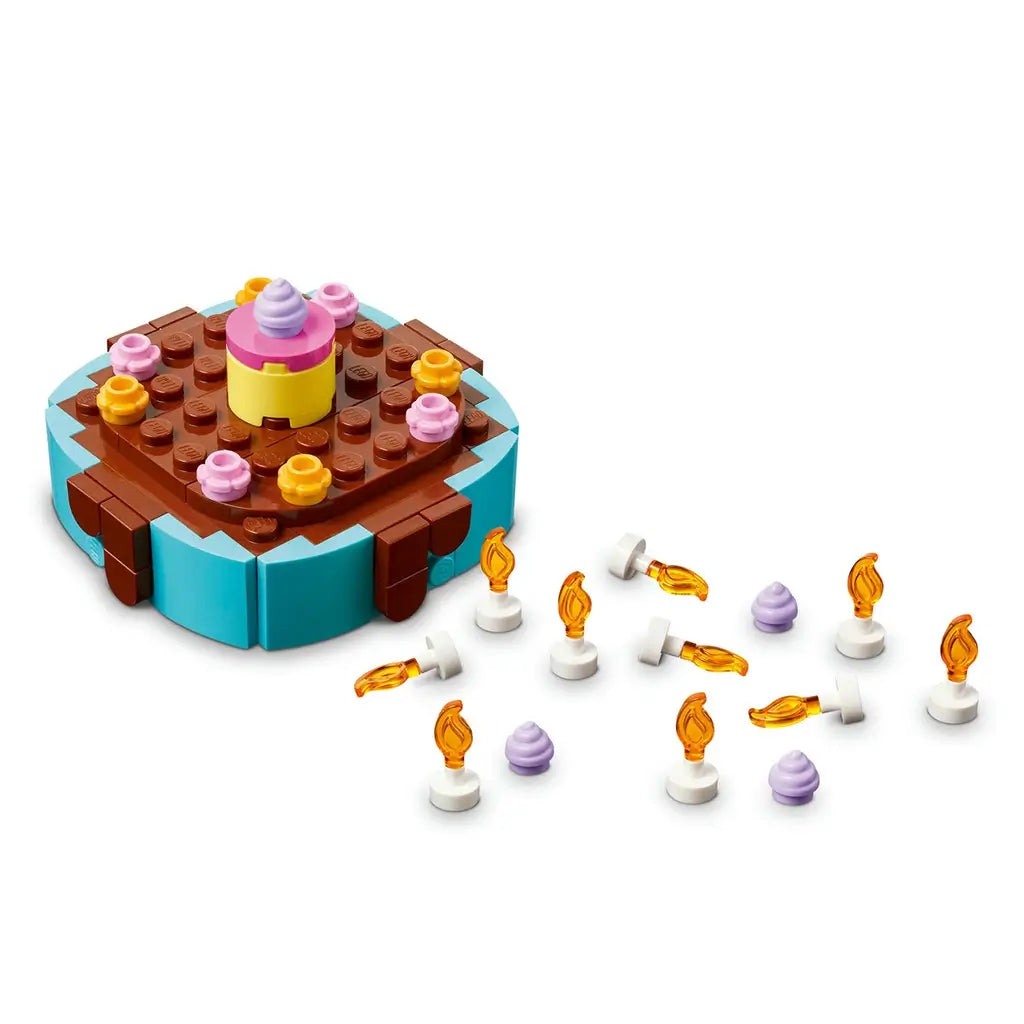 LEGO birthday cake decoration components in blue, brown, pink and yellow plastic bricks, showing a removable cake topper, candle pieces and frosting accents arranged on a plain studio background.