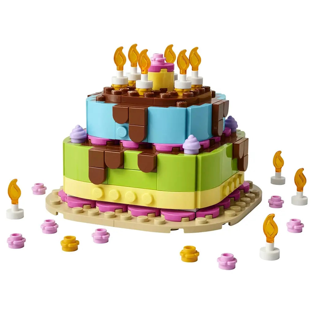LEGO birthday cake display built from blue, green, pink, tan and brown plastic bricks, forming a two-tier celebration cake with candle decorations, frosting accents and party pieces on a plain studio background.