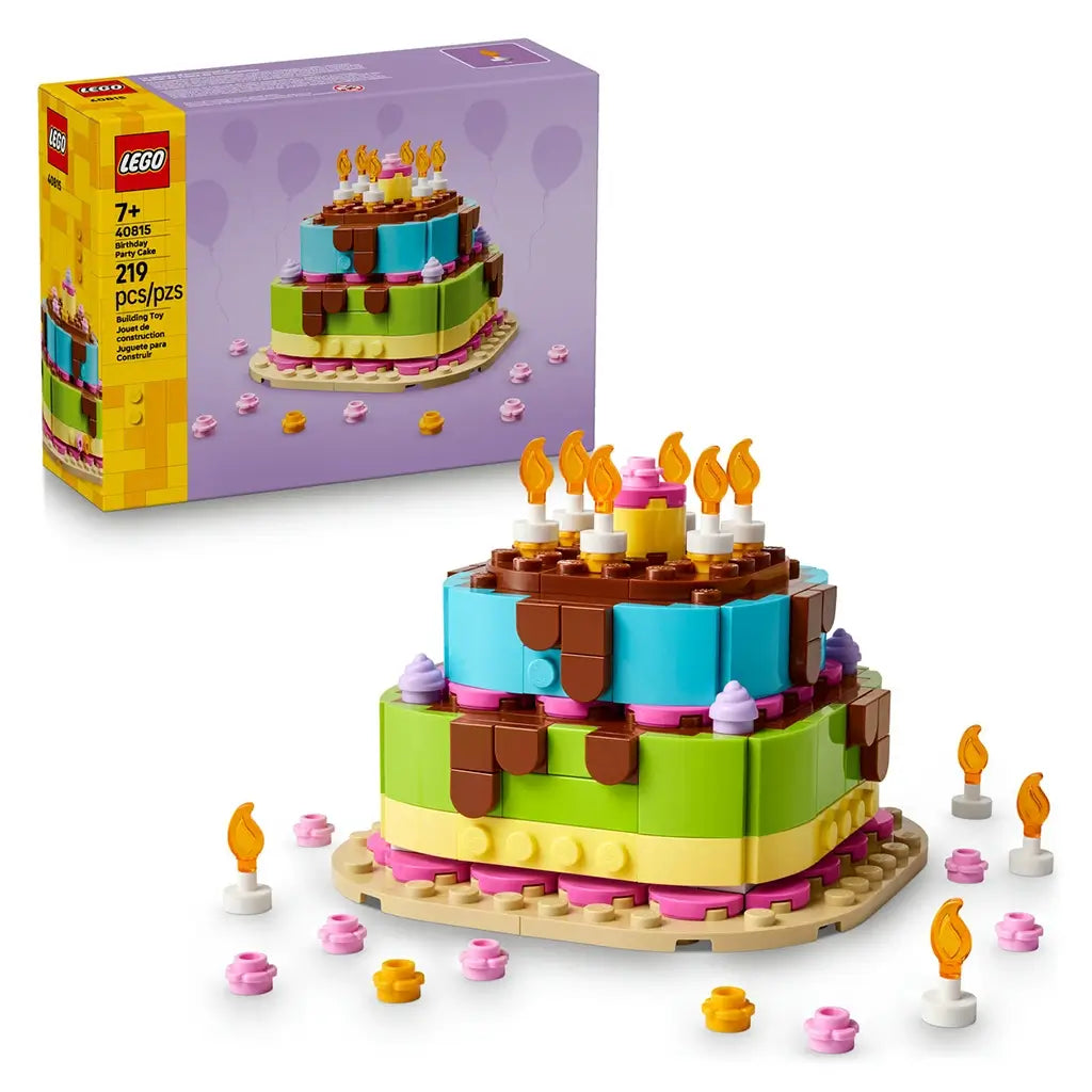 LEGO birthday cake gift set featuring yellow and purple cardboard packaging beside a colorful two-tier plastic brick cake, decorated with candles, frosting details and party elements on a neutral studio background.