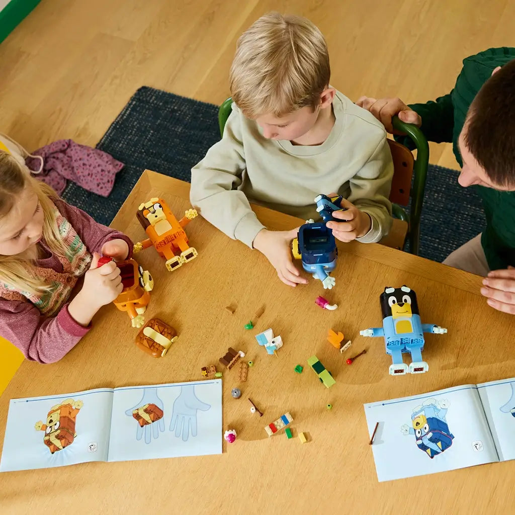 LEGO Bluey Brick-Built Family set in blue, orange, brown, and yellow plastic is assembled by two children at a wooden table, with loose accessories and instructions nearby.