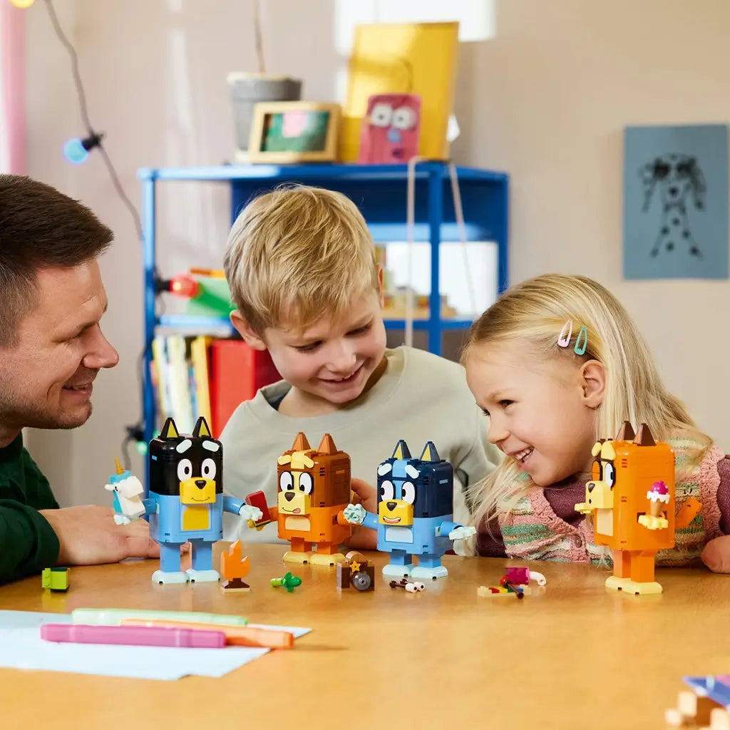 LEGO Bluey Brick-Built Family figures in blue, orange, brown, and yellow plastic stand on a table as an adult and two smiling children play with accessories in a bright room.