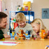 LEGO Bluey Brick-Built Family figures in blue, orange, brown, and yellow plastic stand on a table as an adult and two smiling children play with accessories in a bright room.