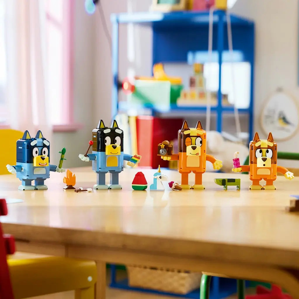 LEGO Bluey Brick-Built Family figures in blue, navy, orange, brown, and yellow plastic stand with colorful accessories across a wooden table in a bright children's playroom.