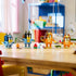 LEGO Bluey Brick-Built Family figures in blue, navy, orange, brown, and yellow plastic stand with colorful accessories across a wooden table in a bright children's playroom.