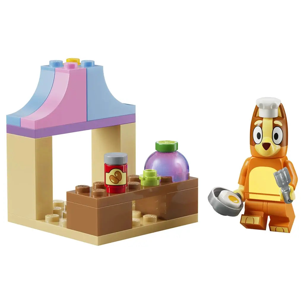 LEGO Bluey restaurant toy in orange, cream, brown, pink and light blue plastic bricks, shaped as a café stand with food items and Bingo chef figure, isolated on a clean studio background.
