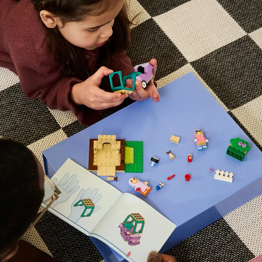 LEGO Bluey Grannies playset in purple, teal, tan, green, pink, and lavender plastic is assembled by a child on a blue surface with loose pieces and illustrated instructions.