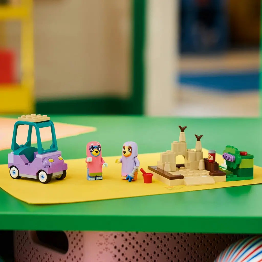 LEGO Bluey Grannies playset in purple, teal, tan, green, pink, and lavender plastic displays a toy car, two figures, sandcastle, bucket, and garden scene on a colorful table.