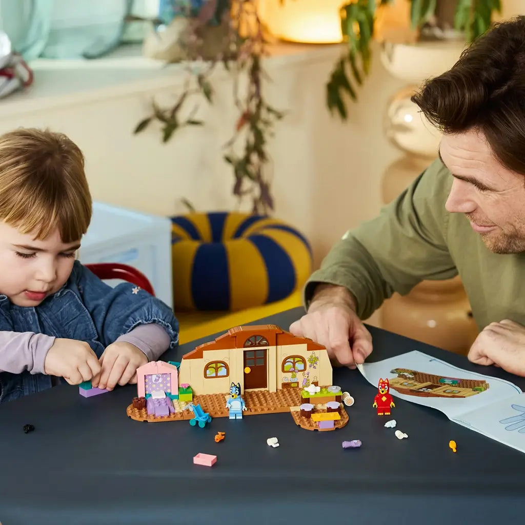LEGO Bluey School set in tan, brown, pink, teal, and purple plastic is assembled on a table by a child and adult, with Bluey, Rusty, accessories, and instructions nearby.