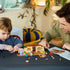 LEGO Bluey School set in tan, brown, pink, teal, and purple plastic is assembled on a table by a child and adult, with Bluey, Rusty, accessories, and instructions nearby.
