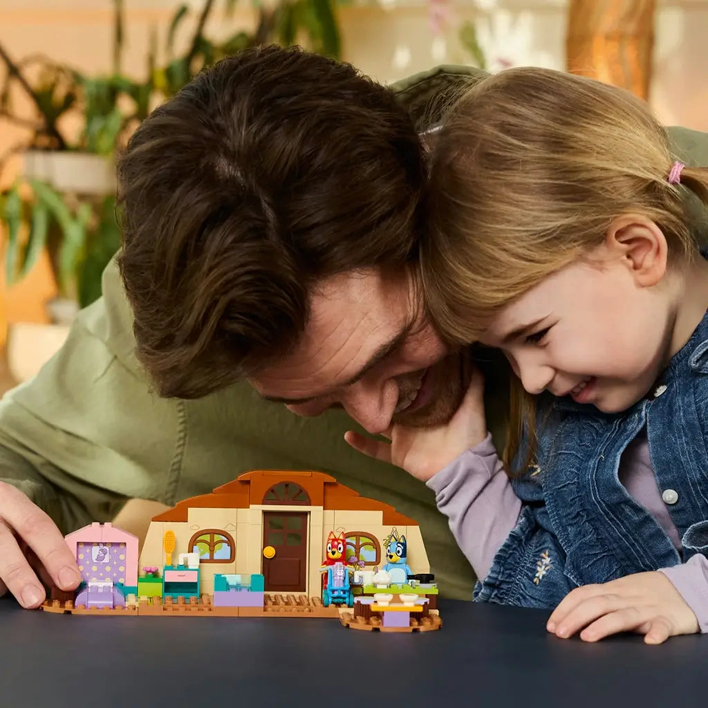 LEGO Bluey School set in tan, brown, pink, and teal plastic sits on a table as an adult and child play with Bluey and Rusty figures in a bright indoor setting.