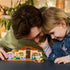 LEGO Bluey School set in tan, brown, pink, and teal plastic sits on a table as an adult and child play with Bluey and Rusty figures in a bright indoor setting.