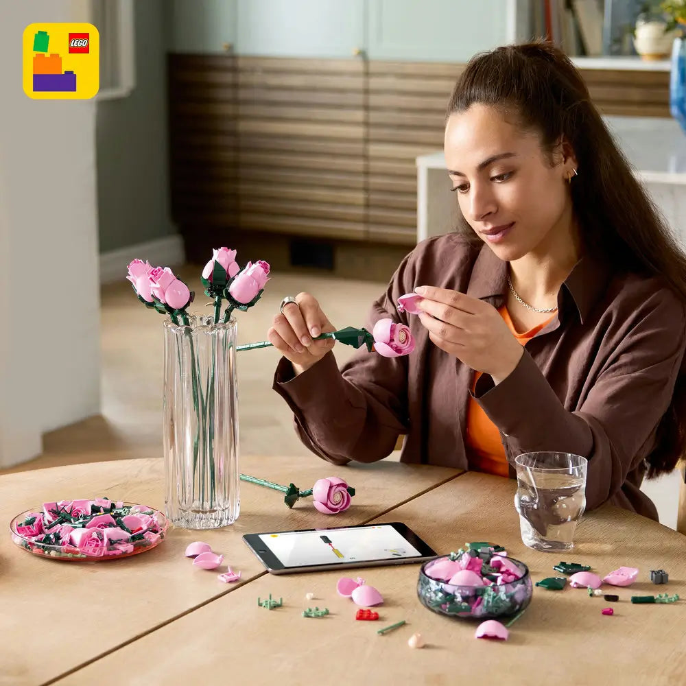A woman assembles LEGO pink roses with glossy plastic petals and green stems, carefully shaping each curved bloom while seated at a wooden table in a bright home setting. Designed for detailed display with realistic plastic floral elements.